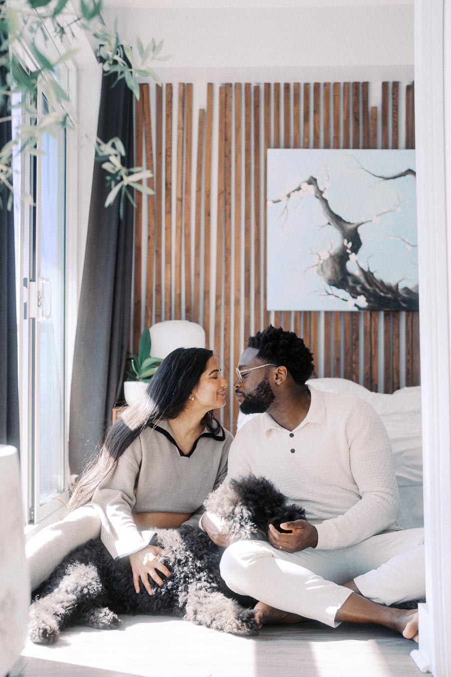 Couple sitting together in a cozy bedroom with a black fluffy dog, wooden slat wall background, and abstract tree painting.