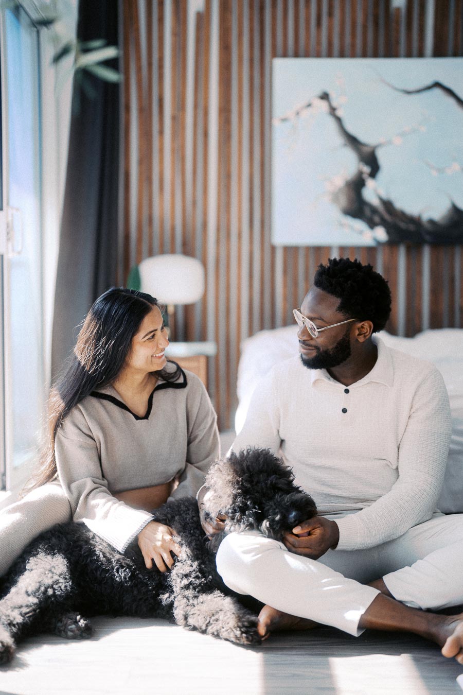 Couple sitting on the floor, smiling and petting a black poodle in a modern living room with natural light.