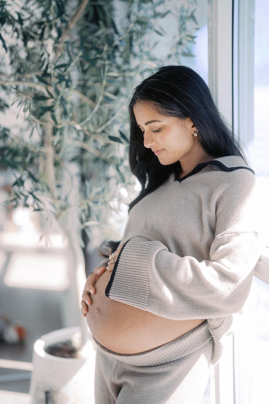 Pregnant woman in cozy sweater lovingly cradles her belly by a sunlit window, with a green plant in the background, conveying warmth and anticipation.