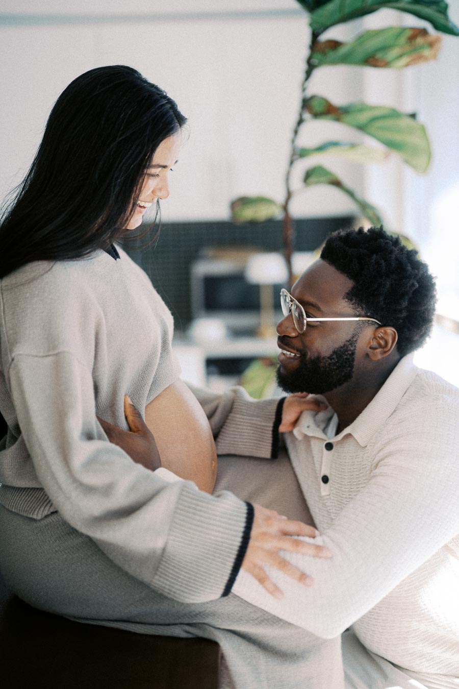 A joyful couple embracing during a maternity moment, with the expectant mother showing her baby bump while the father smiles lovingly. The warm home setting and casual attire create a cozy and intimate atmosphere.