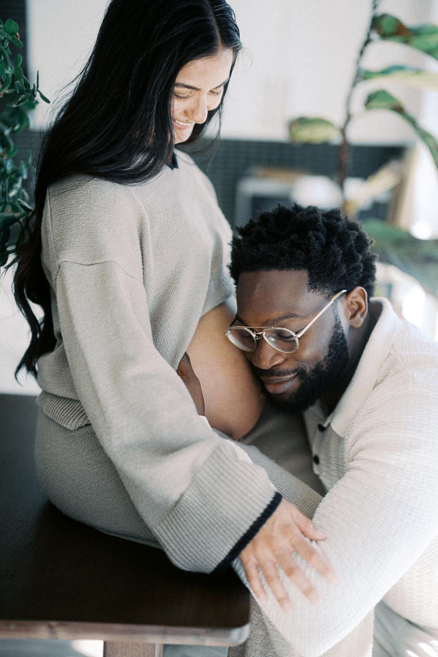 Happy couple embracing during pregnancy, woman sitting on a table, and man gently resting his head on her belly, capturing a tender moment of anticipation and love in a cozy indoor setting.