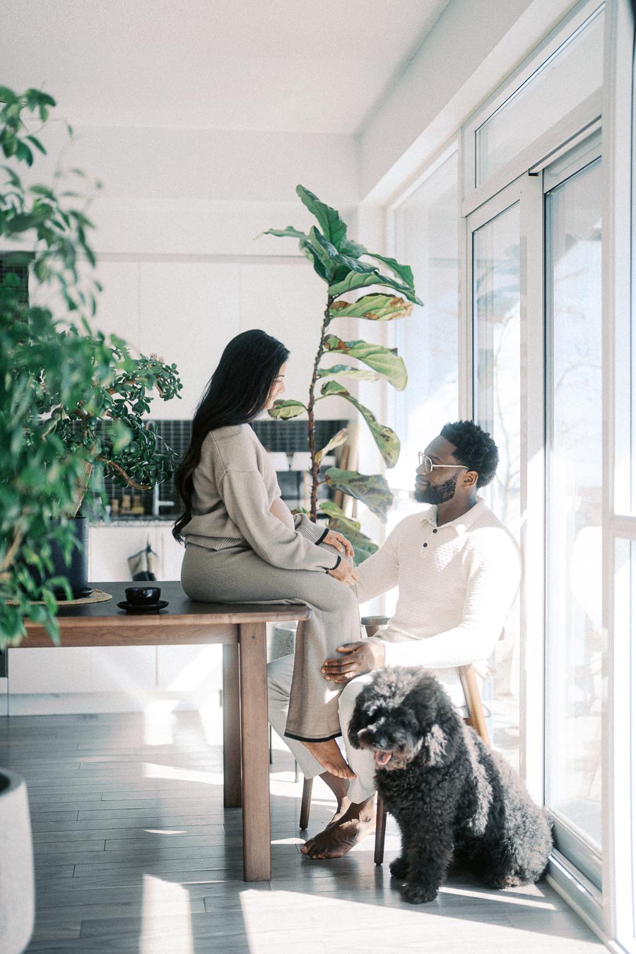 A couple enjoying a peaceful moment in a sunlit room with large houseplants, sitting at a wooden table near a window. A fluffy black dog sits contentedly beside them, enhancing the cozy and serene atmosphere of the modern home.