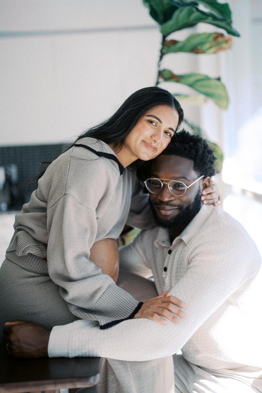 A happy couple embracing in a cozy indoor setting with natural light and plants in the background.