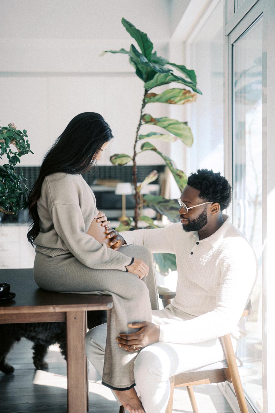 Couple sharing a tender moment during pregnancy, indoors by a large sunny window with lush green plants in the background. The woman is seated on a table as the man gently touches her baby bump, capturing an intimate and joyful stage of parenthood.