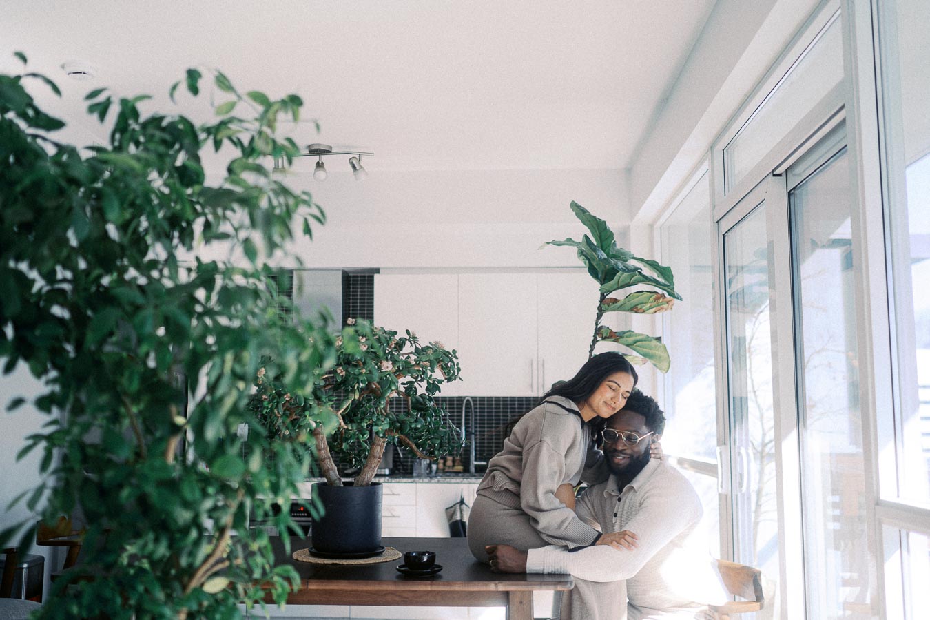 A couple sharing a warm embrace in a modern, sunlit living room with lush green indoor plants and large windows in the background.