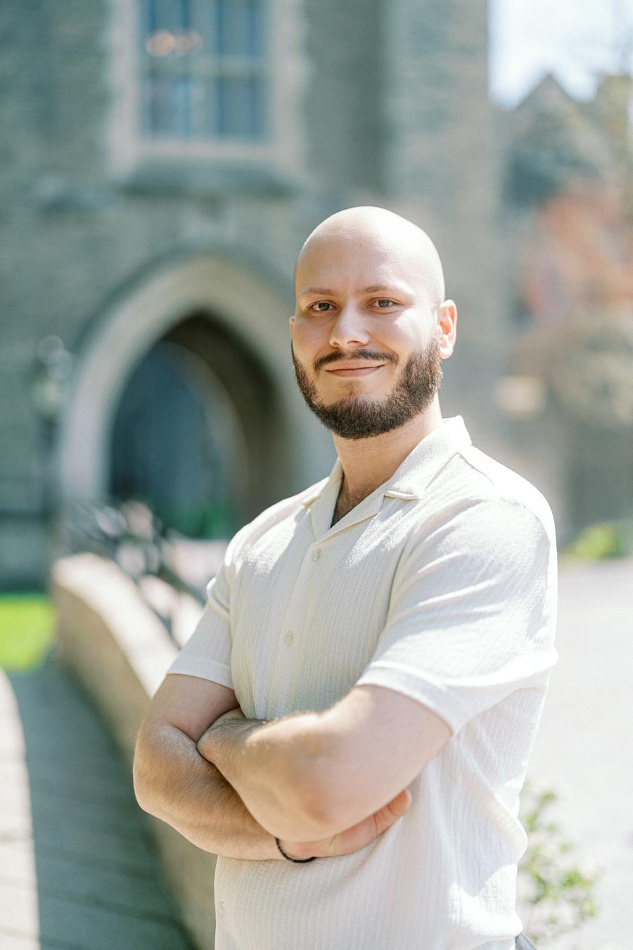 Bald man with a beard smiling confidently, arms crossed, standing in sunlight near a stone archway at the University of Toronto.