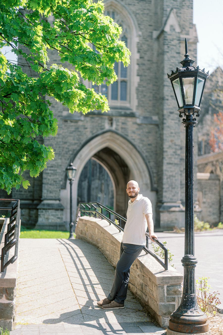 Man standing by a stone railing near a gothic archway and vintage lamp post at University of Toronto, surrounded by bright green spring leaves.