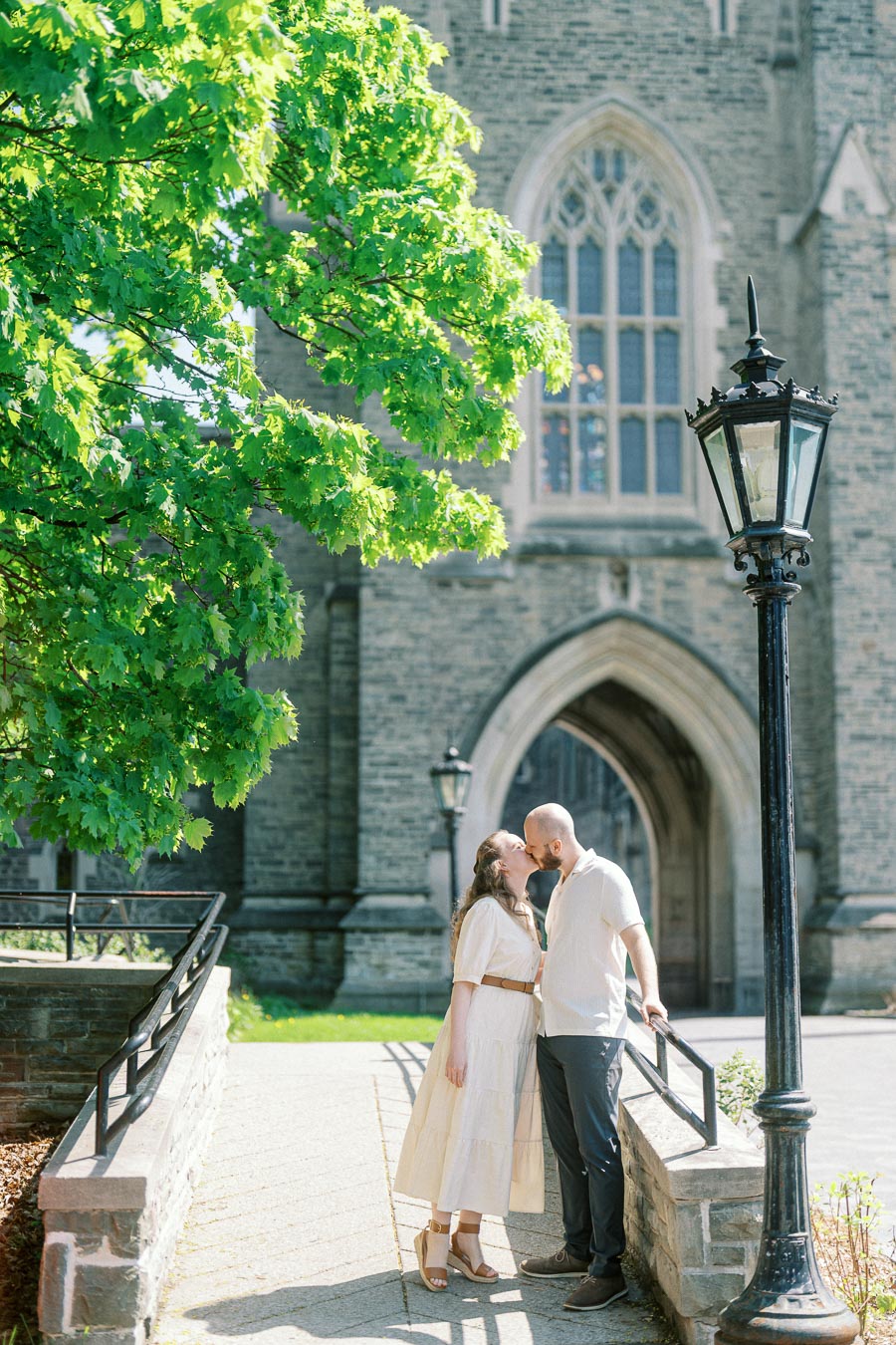 Couple shares a romantic kiss near a stone building and blooming green tree at University of Toronto on a sunny spring day.
