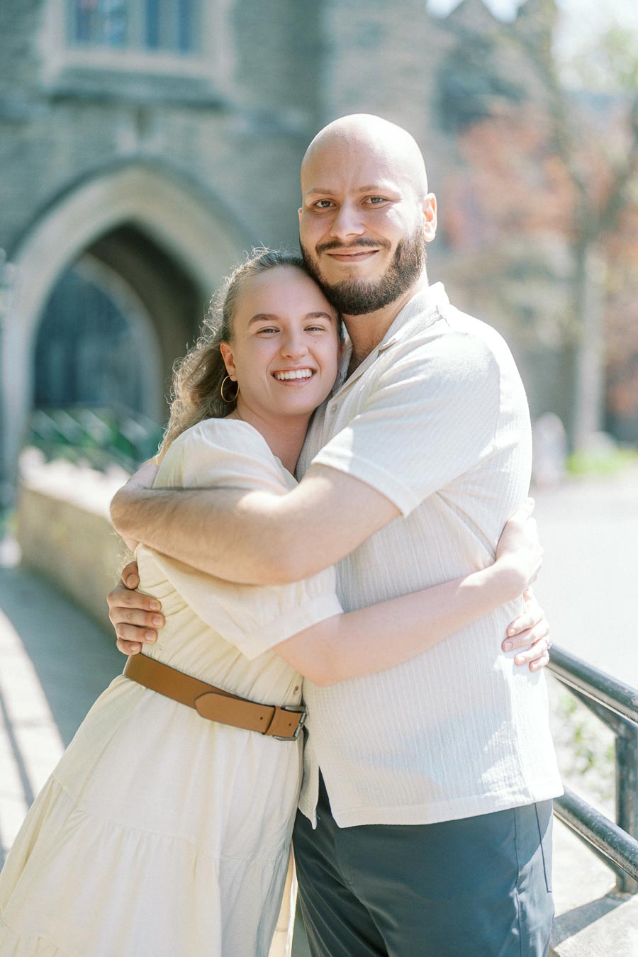 Happy engaged couple hugging and smiling outdoors in spring at the University of Toronto, with historic stone architecture in the background.