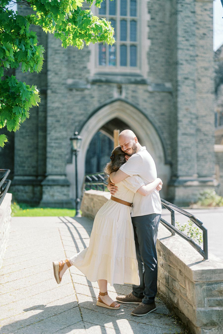 Couple hugging warmly in front of a stone archway at University of Toronto on a sunny spring day, surrounded by greenery.