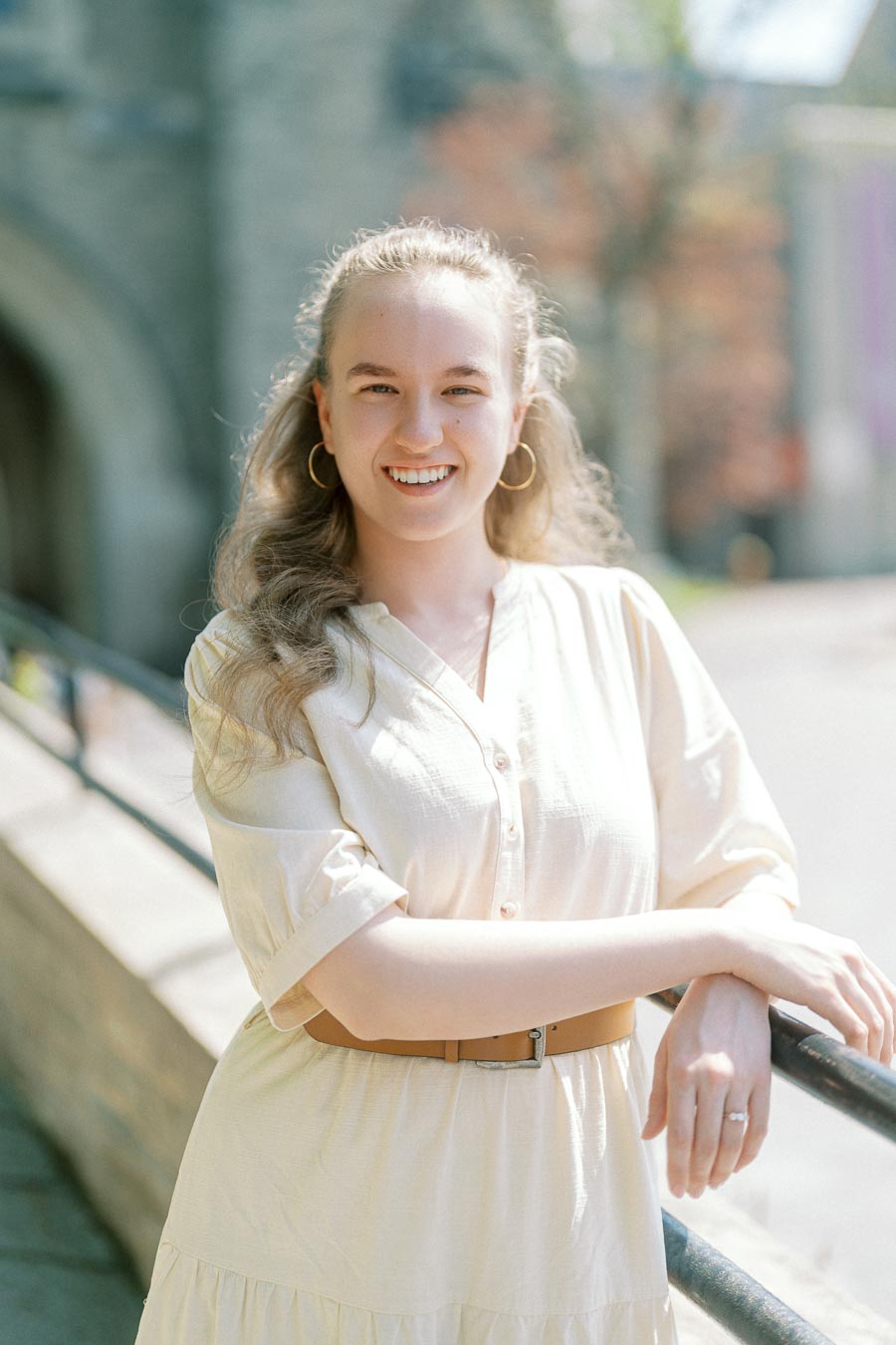 Woman in a light dress smiling and leaning on a railing with University of Toronto's historic architecture in the background.