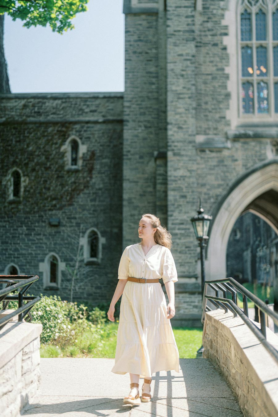 Woman in a cream dress walking along a sunlit path beside historic stone buildings at the University of Toronto in spring.