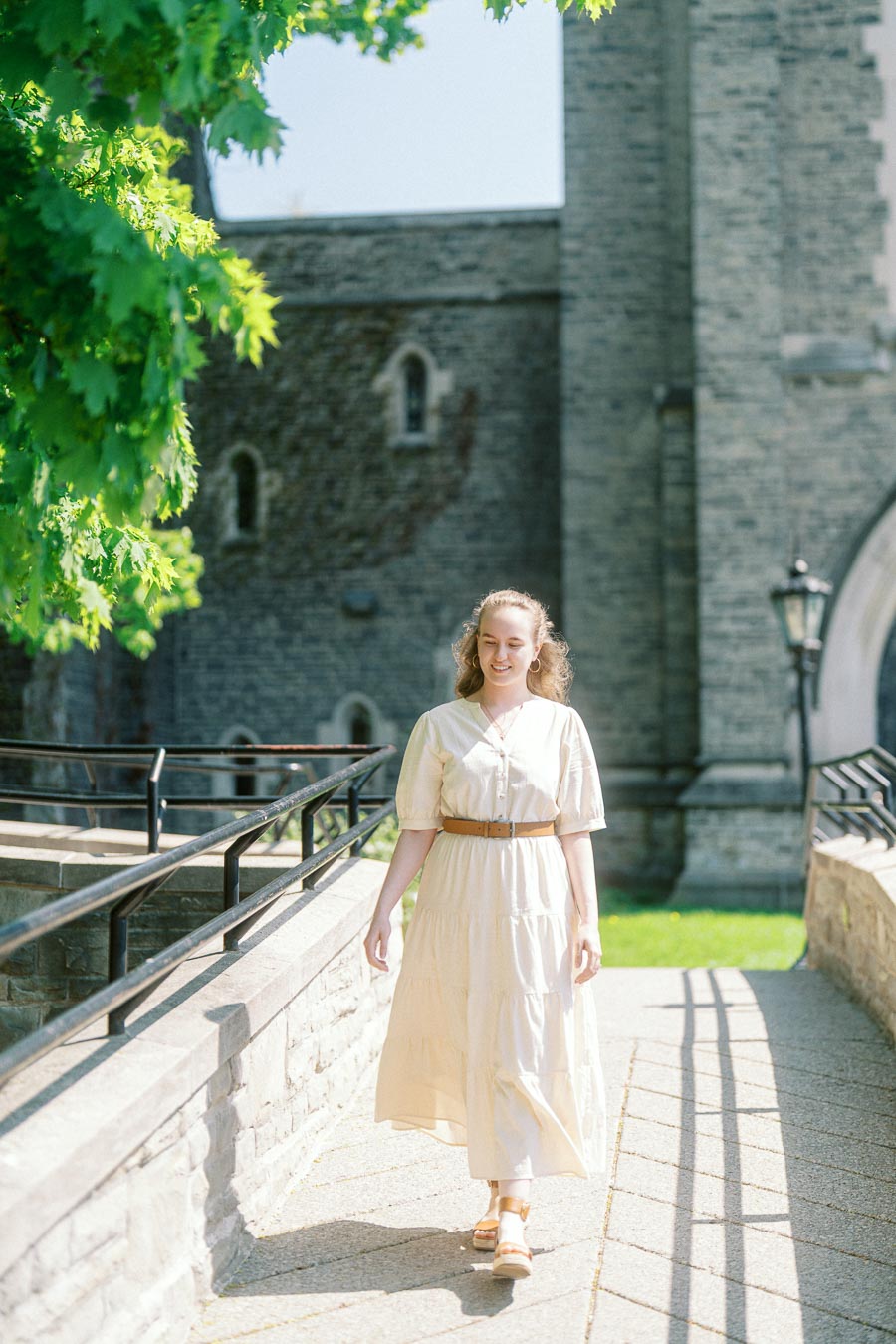 Woman in a light dress walks along a sunlit stone path beside historic University of Toronto architecture and lush spring greenery.