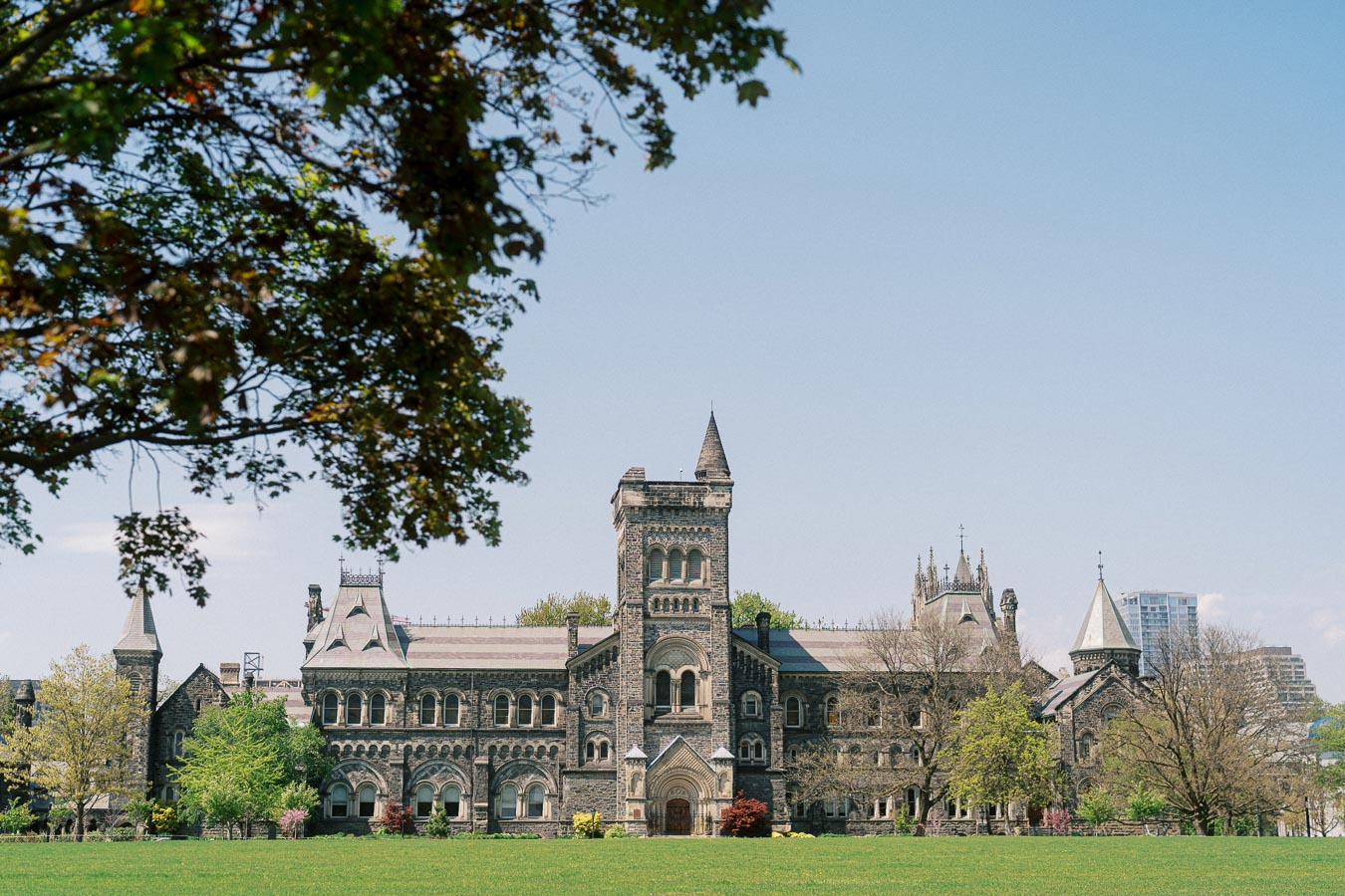 University of Toronto's historic stone building framed by leafy trees and a green lawn on a sunny spring day.