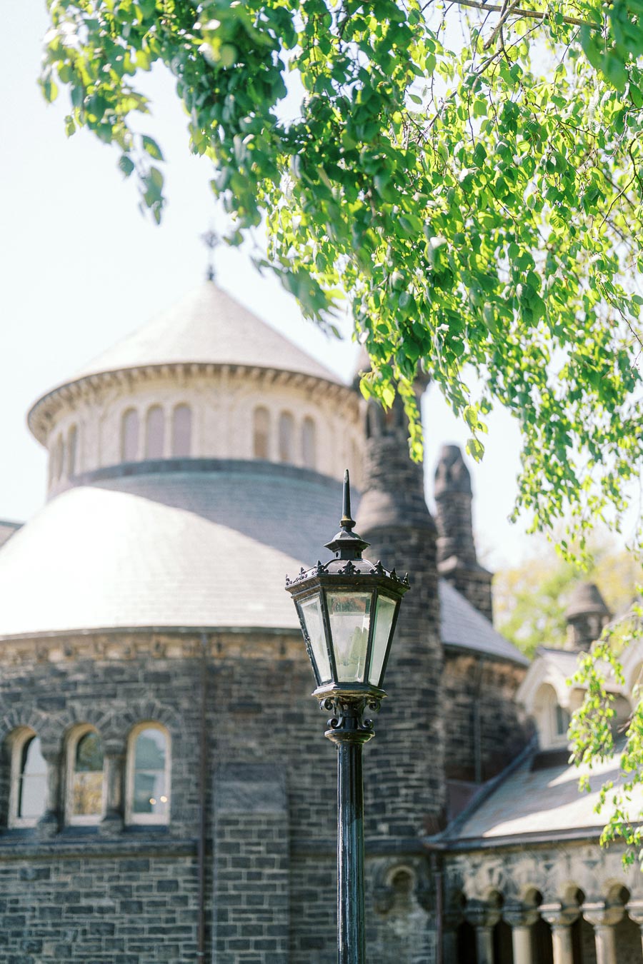 Vintage lamp post in front of University of Toronto’s round stone building and green spring leaves overhead.