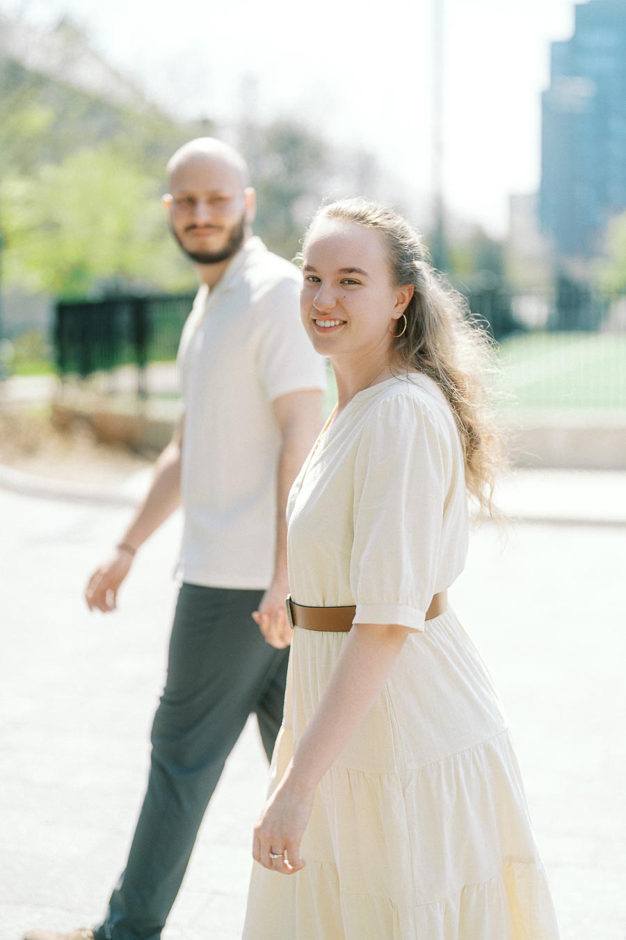 Smiling engaged couple walking hand in hand in soft spring sunlight at University of Toronto, with greenery and campus buildings in the background.