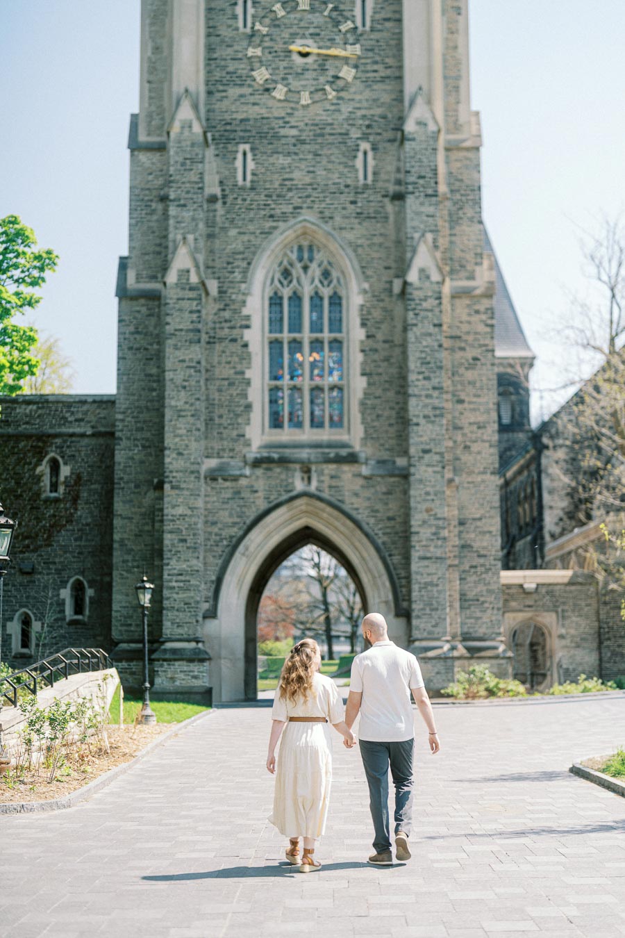 A couple walks hand-in-hand toward a grand stone archway at the University of Toronto on a sunny spring day, surrounded by greenery.