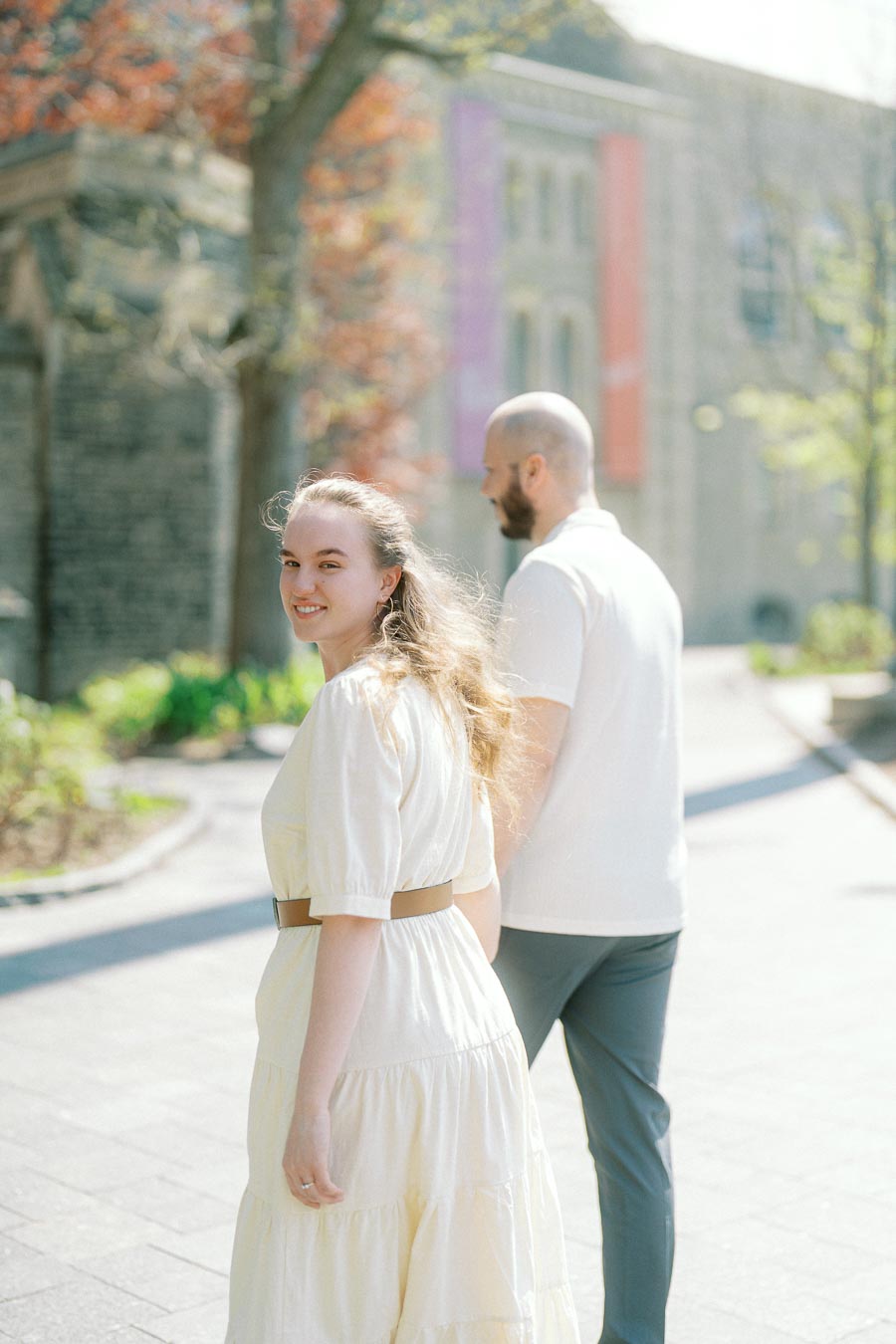 Engaged couple walking on a bright spring day at the University of Toronto, with the woman smiling back and campus architecture in the background.