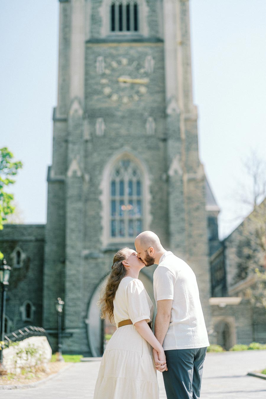 Couple kisses in front of a historic stone clock tower at University of Toronto, holding hands on a sunny spring day.