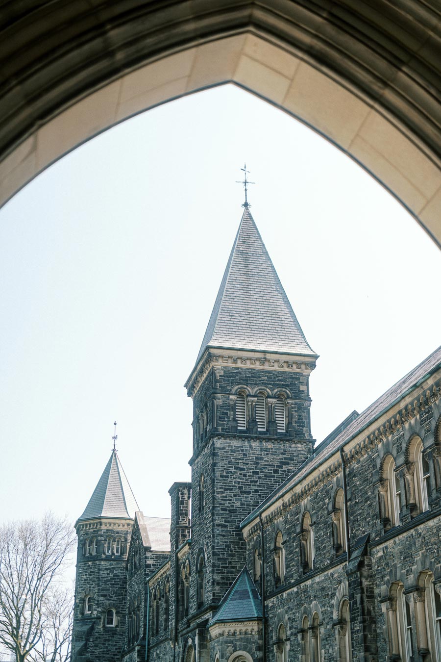 University of Toronto's historic stone tower framed by a stone arch, with clear spring sky and bare trees in the background.