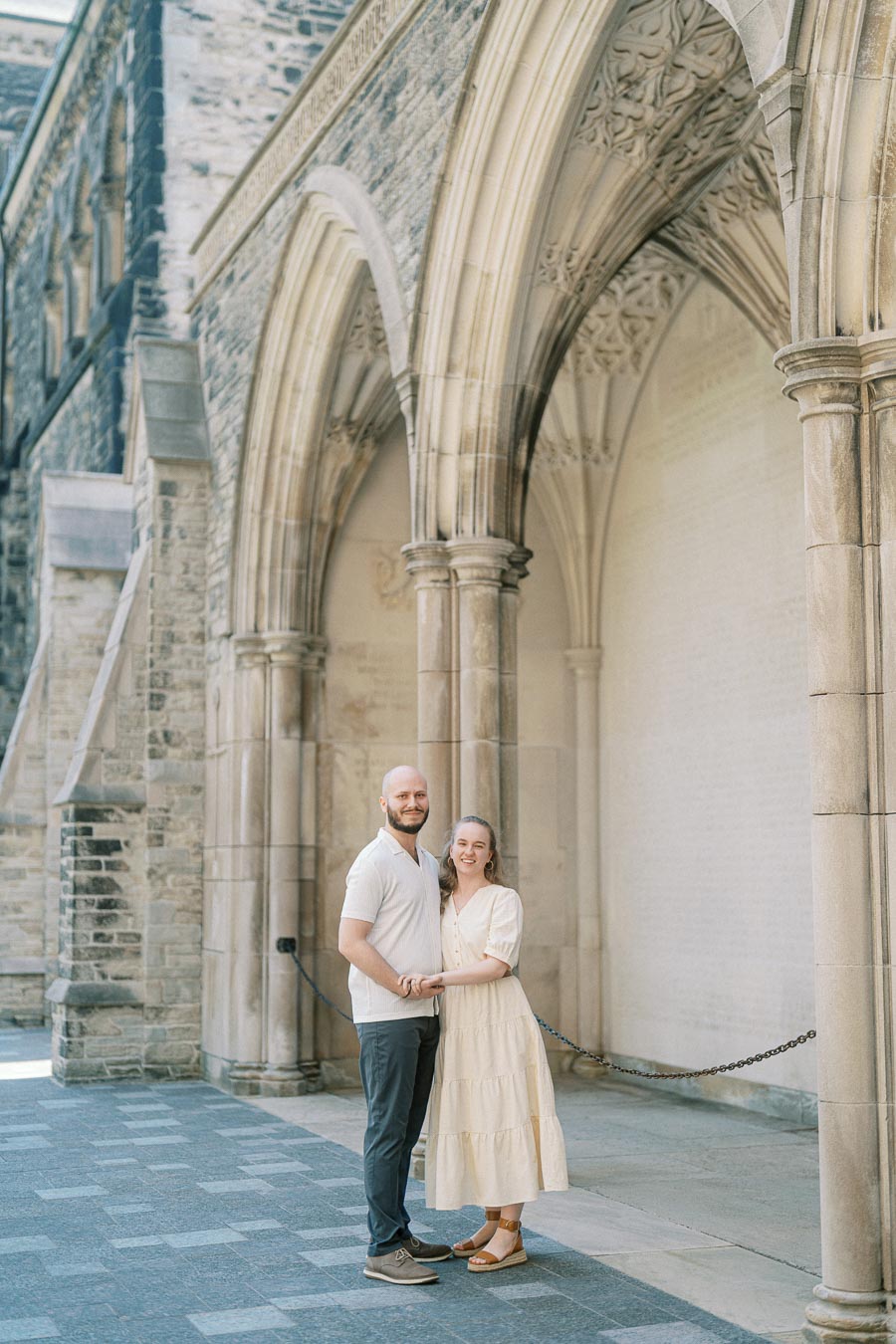 Engaged couple holding hands and smiling beneath grand stone arches at the University of Toronto on a sunny spring day.