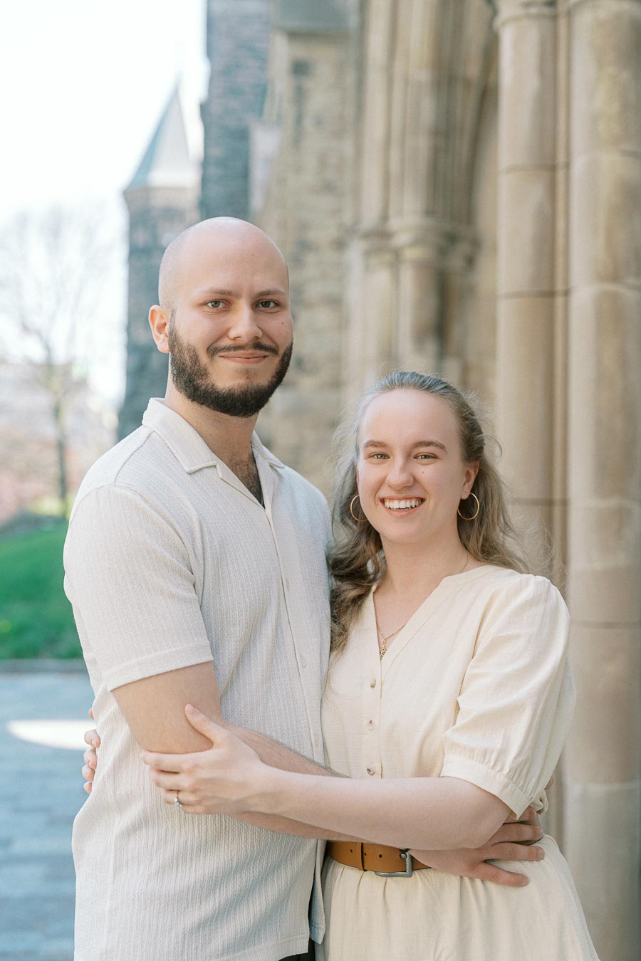 Engaged couple smiling and embracing by a stone archway at the University of Toronto on a sunny spring day.