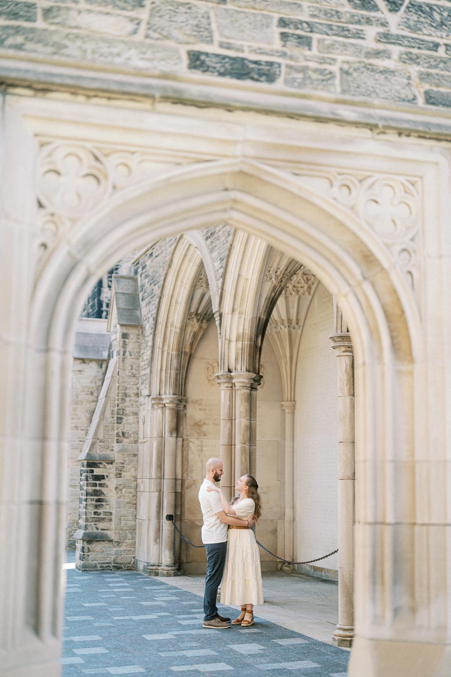 A couple stands embracing under ornate stone arches at the University of Toronto, surrounded by historic architecture and soft spring light.