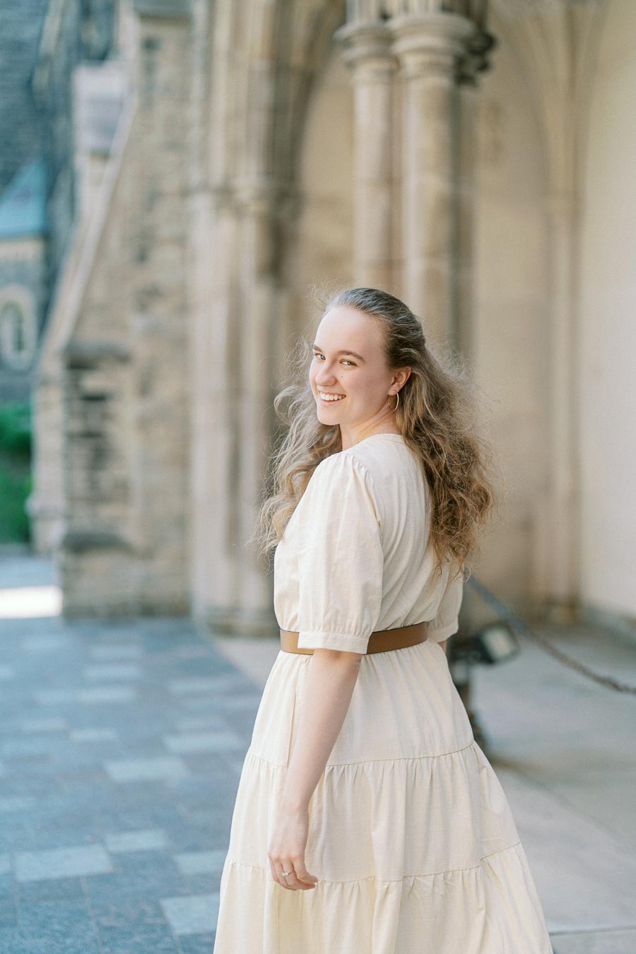 Woman in a cream dress smiling over her shoulder, standing by historic stone arches at the University of Toronto on a sunny spring day.