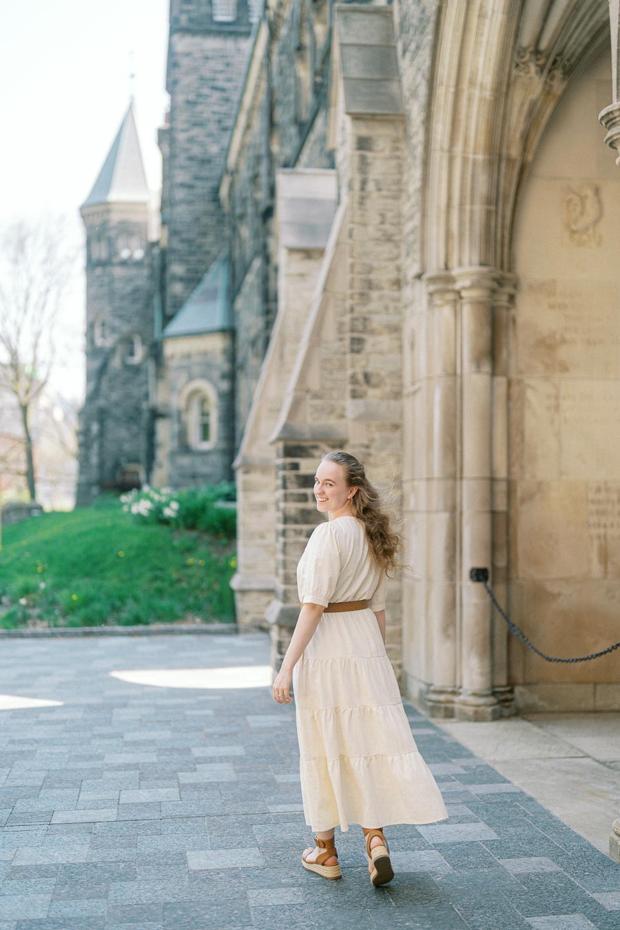 Woman in a cream dress smiles while walking under a stone archway at the University of Toronto on a sunny spring day.
