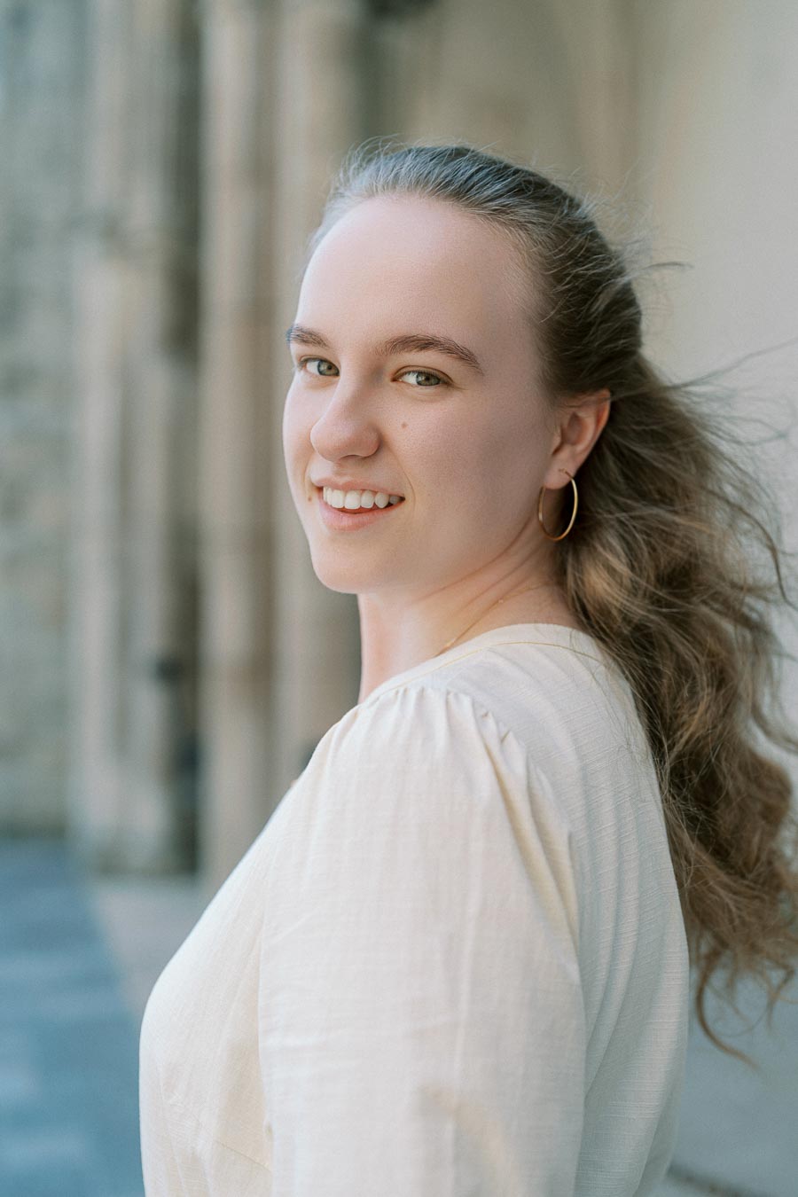Woman with light brown hair smiles in natural light near a stone archway at the University of Toronto during a spring engagement session.