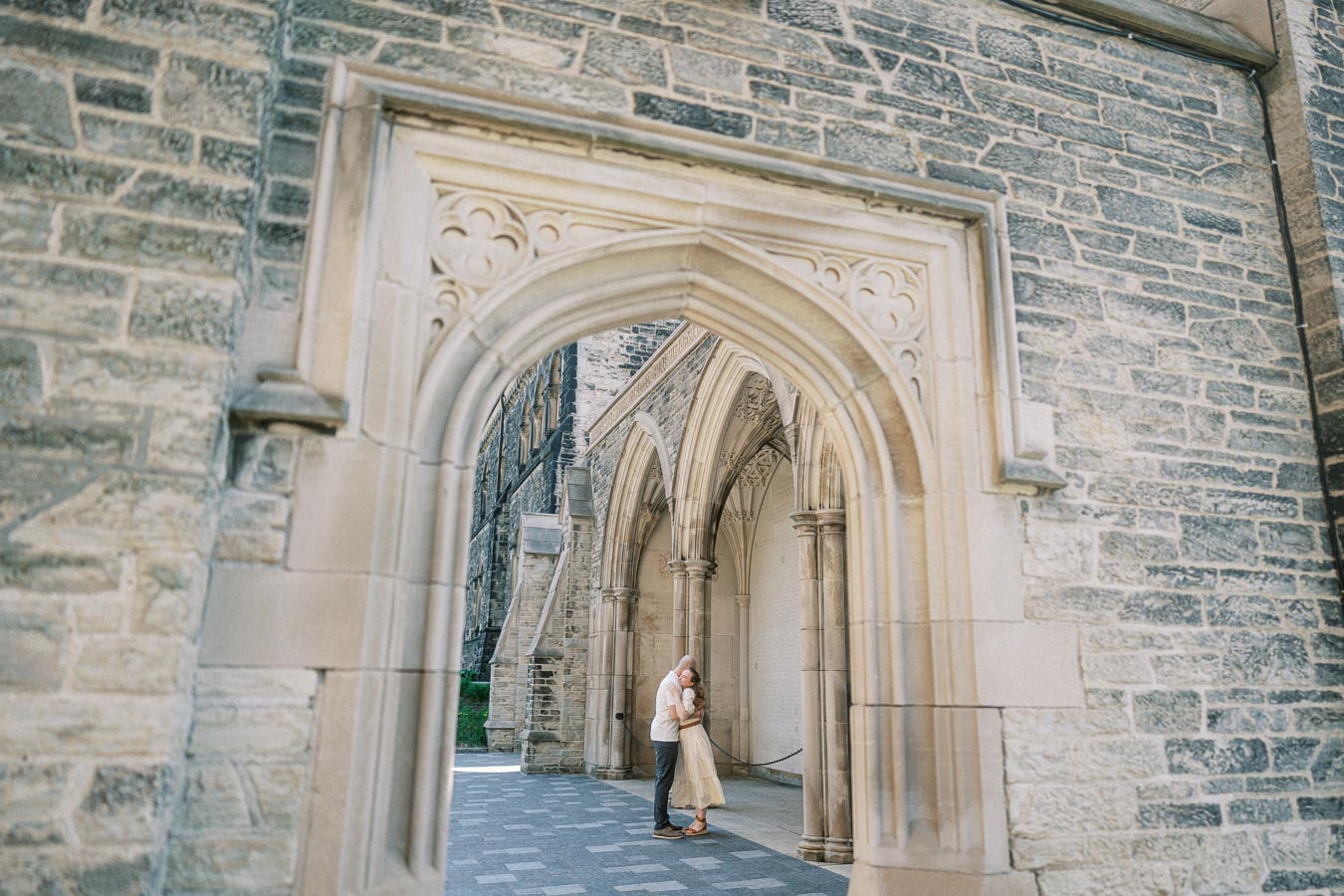 Couple embraces under an ornate stone archway at University of Toronto, surrounded by historic architecture and soft spring daylight.