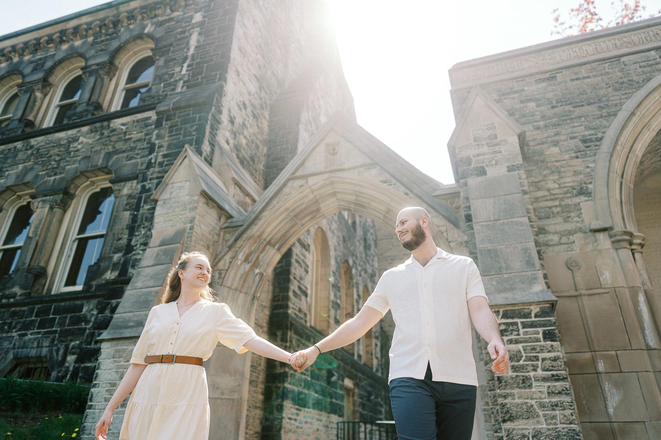 Engaged couple holding hands and smiling under a sunlit stone archway at the University of Toronto, surrounded by historic campus architecture.