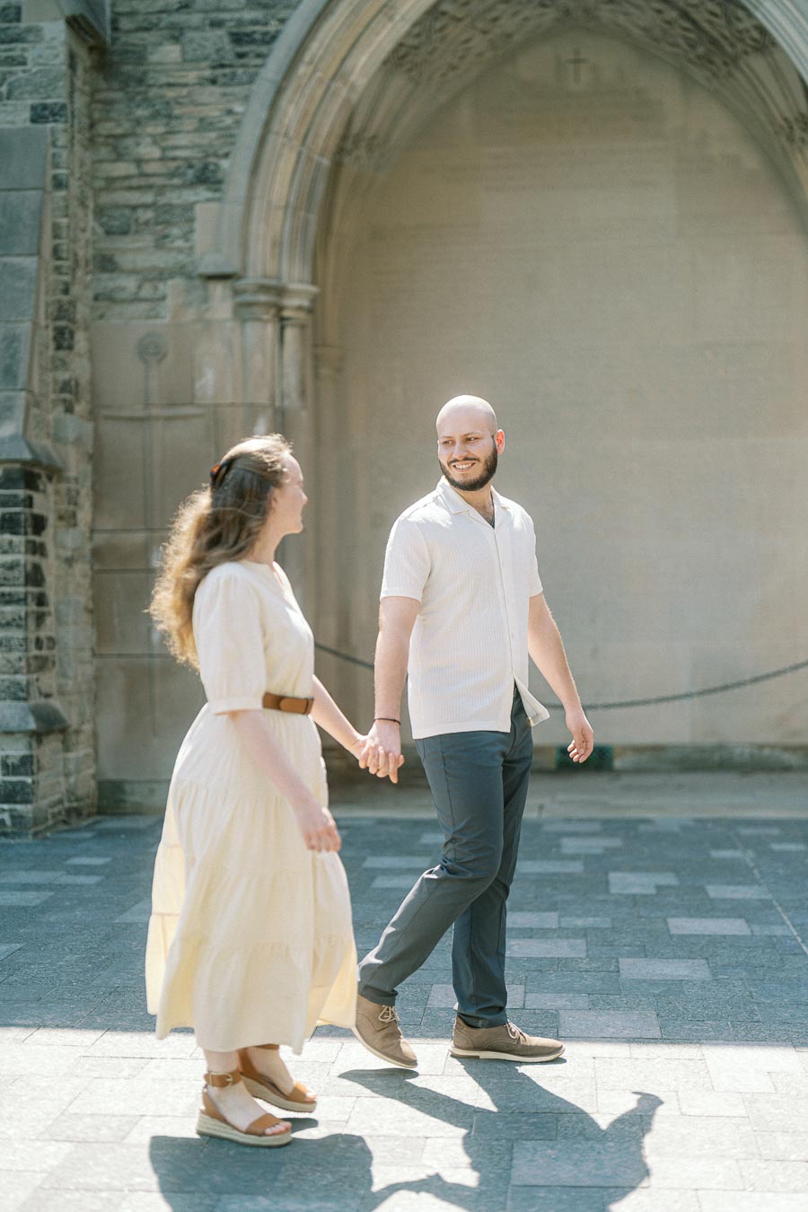 Couple holding hands and smiling while walking in sunlight under a stone archway at the University of Toronto during their spring engagement session.