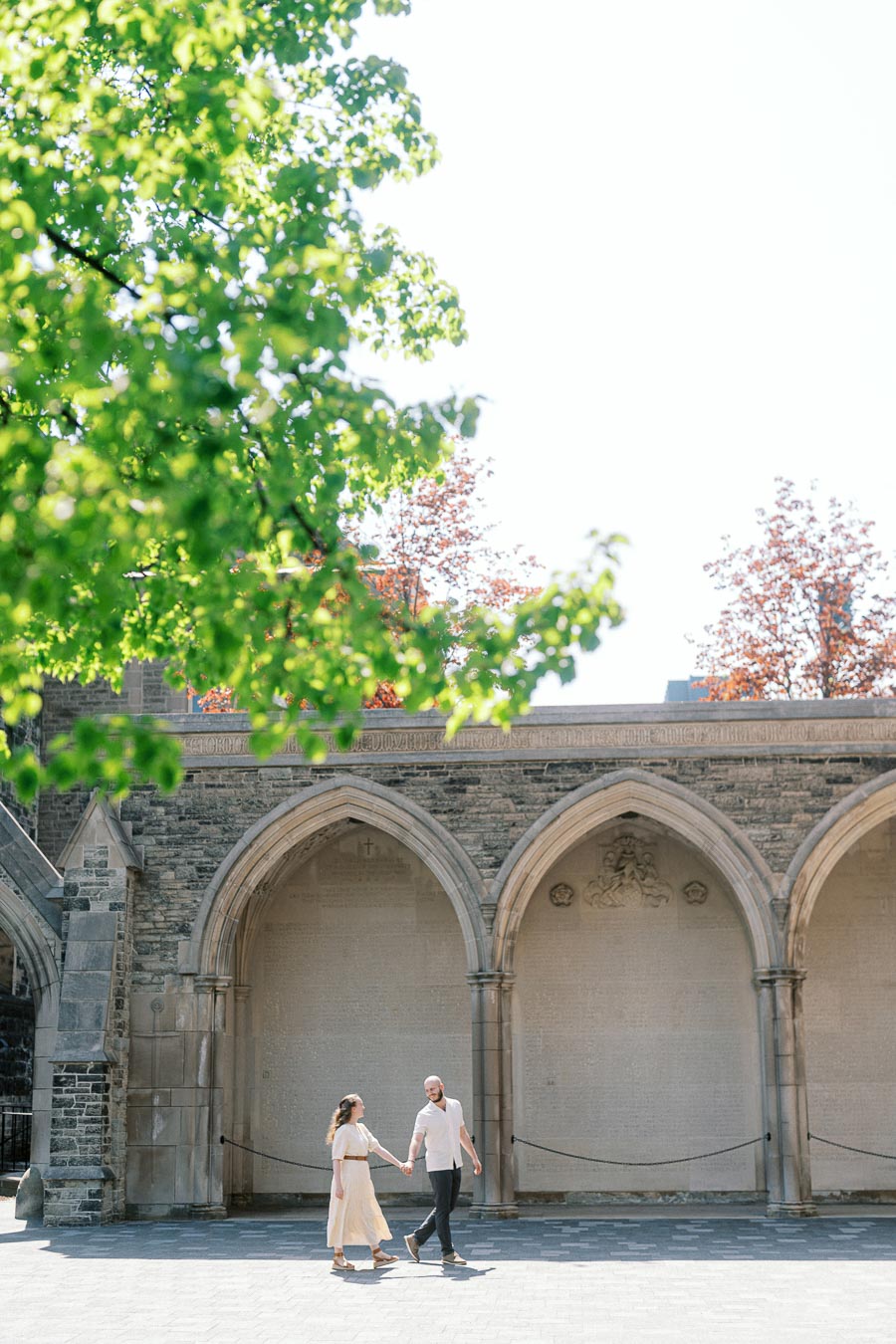 Couple holding hands and walking under historic stone arches at University of Toronto courtyard, with lush green trees and bright spring sunlight.