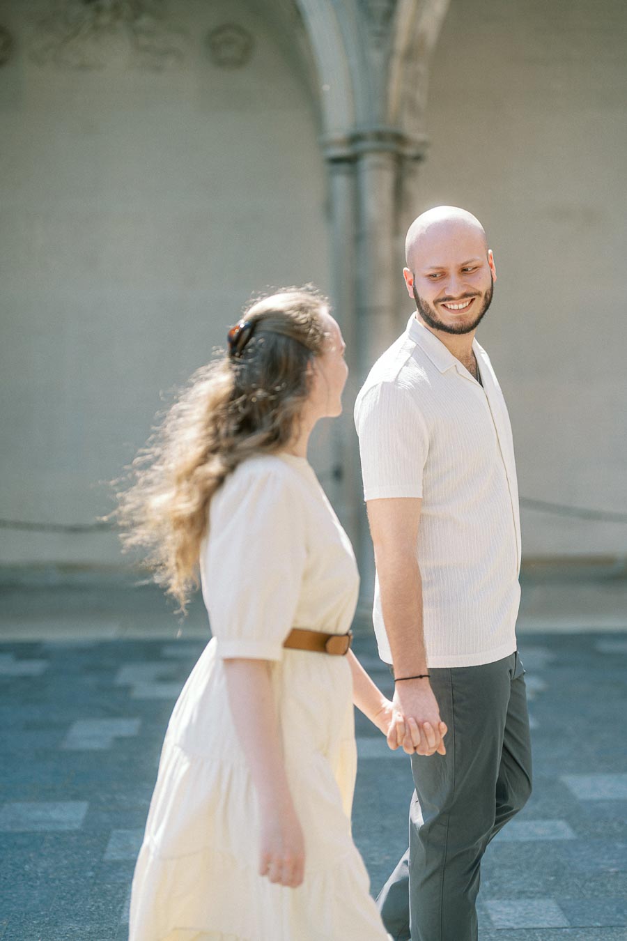Couple holding hands and smiling at each other in a sunlit courtyard at the University of Toronto, surrounded by historic architecture.