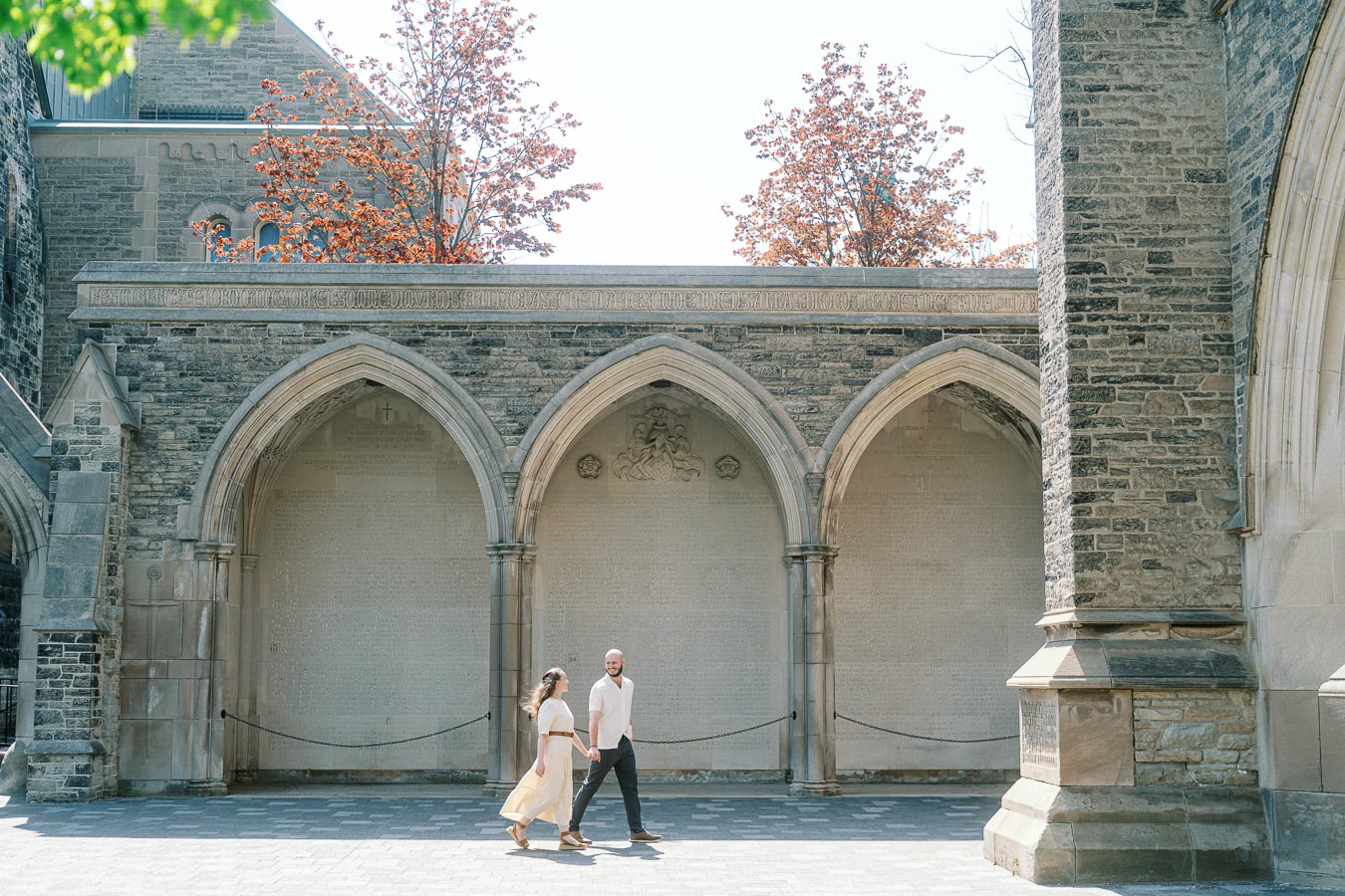 Couple walks hand in hand beneath historic stone arches at University of Toronto, surrounded by spring trees and soft sunlight.