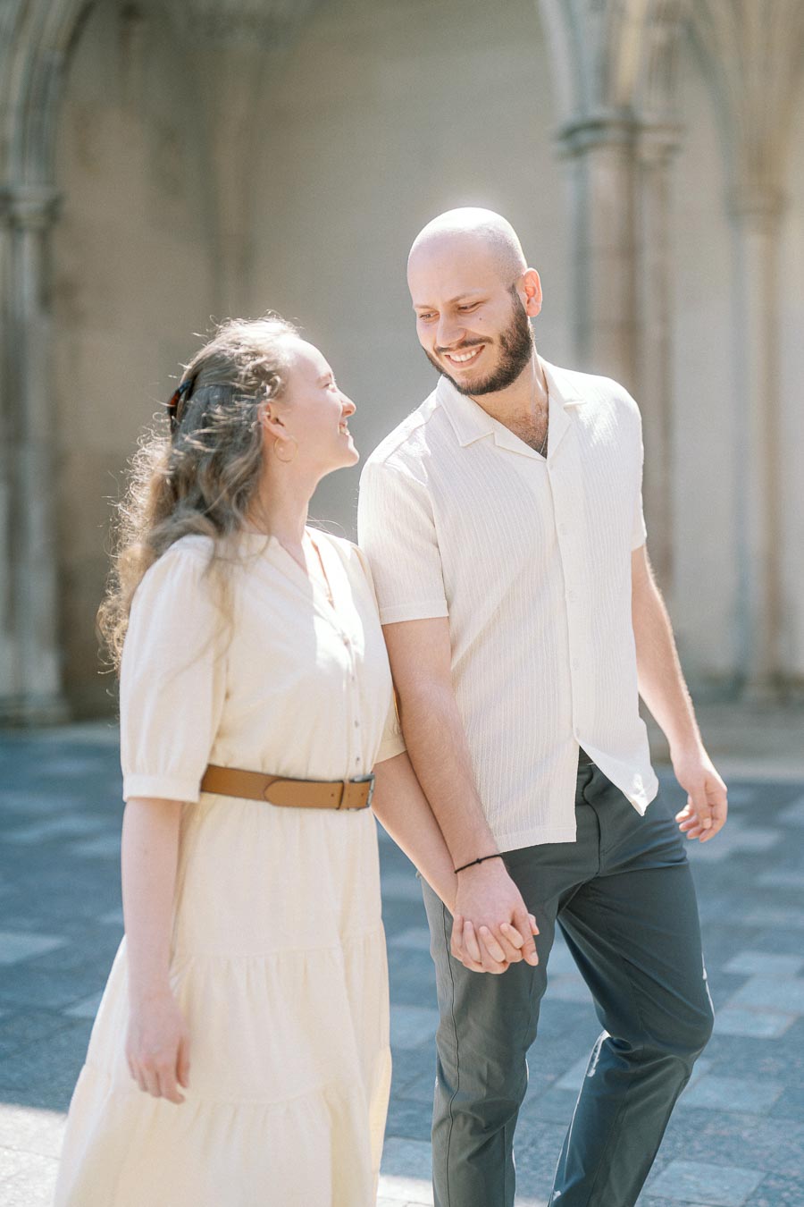 Engaged couple smiling and holding hands while walking beneath sunlit stone arches at University of Toronto in spring.