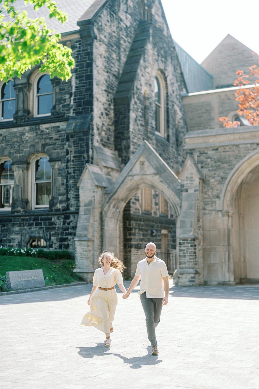 Engaged couple walks joyfully in sunlight by historic stone arches on the University of Toronto campus during spring.