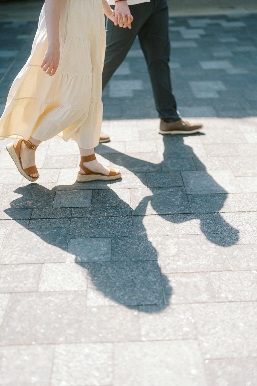 Couple holding hands and walking on a sunny stone path, casting long shadows during a spring engagement session at University of Toronto.