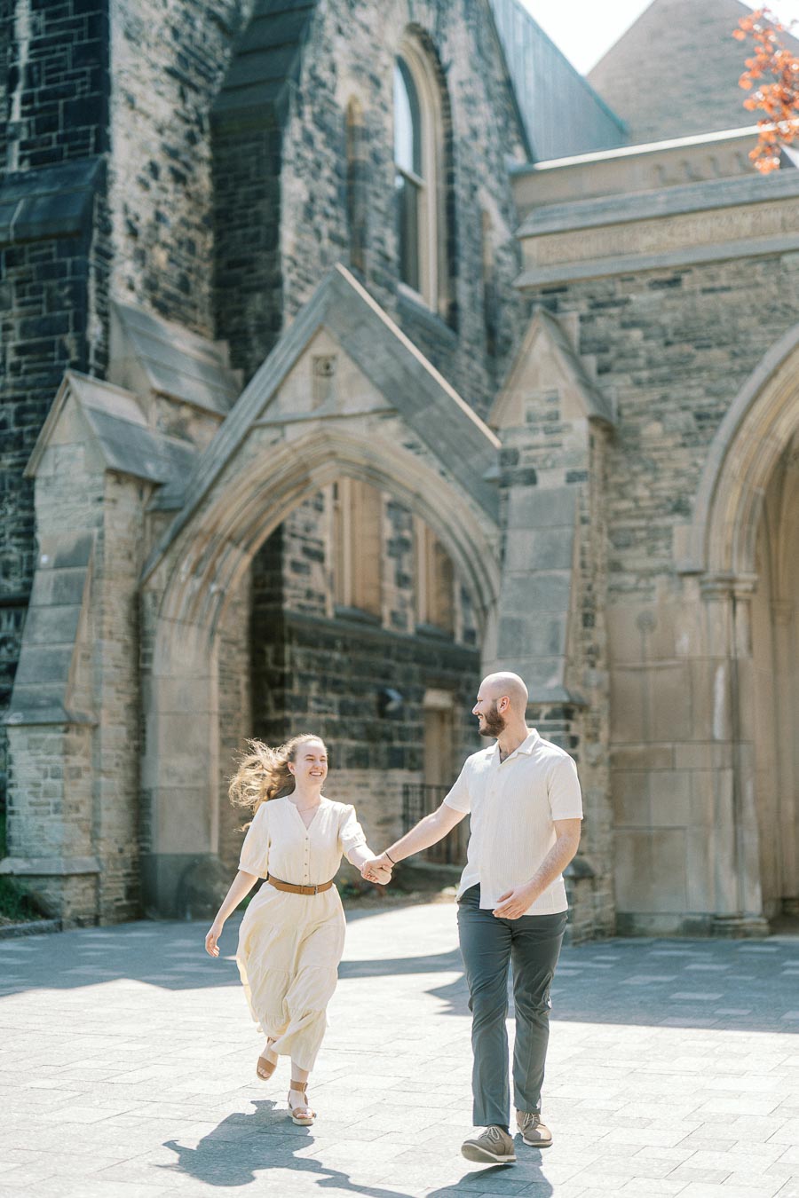 Engaged couple holding hands and walking joyfully in front of stone arches at the University of Toronto on a sunny spring day.