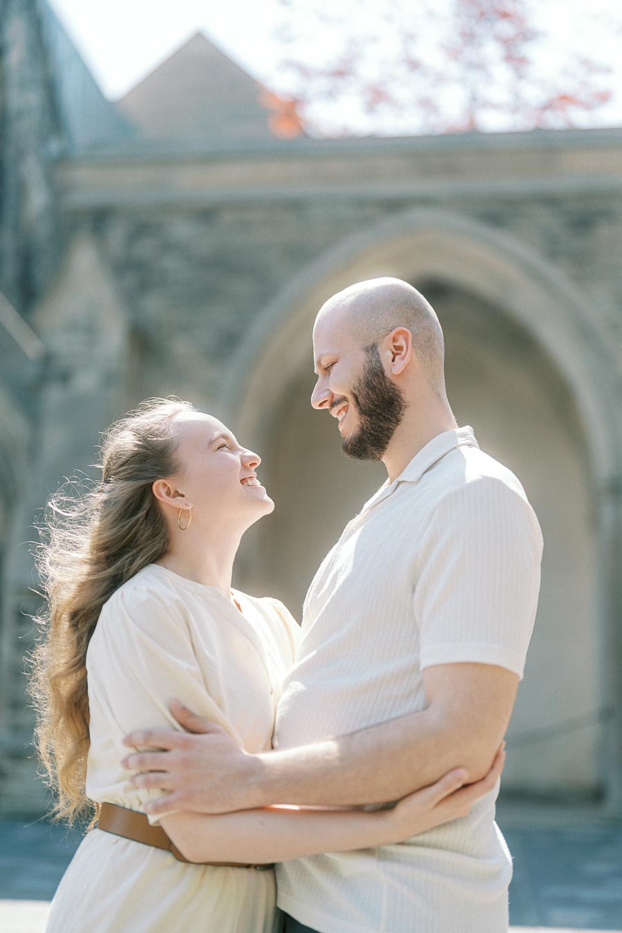 Engaged couple smiling at each other in a warm embrace beneath a stone archway at the University of Toronto on a sunny spring day.
