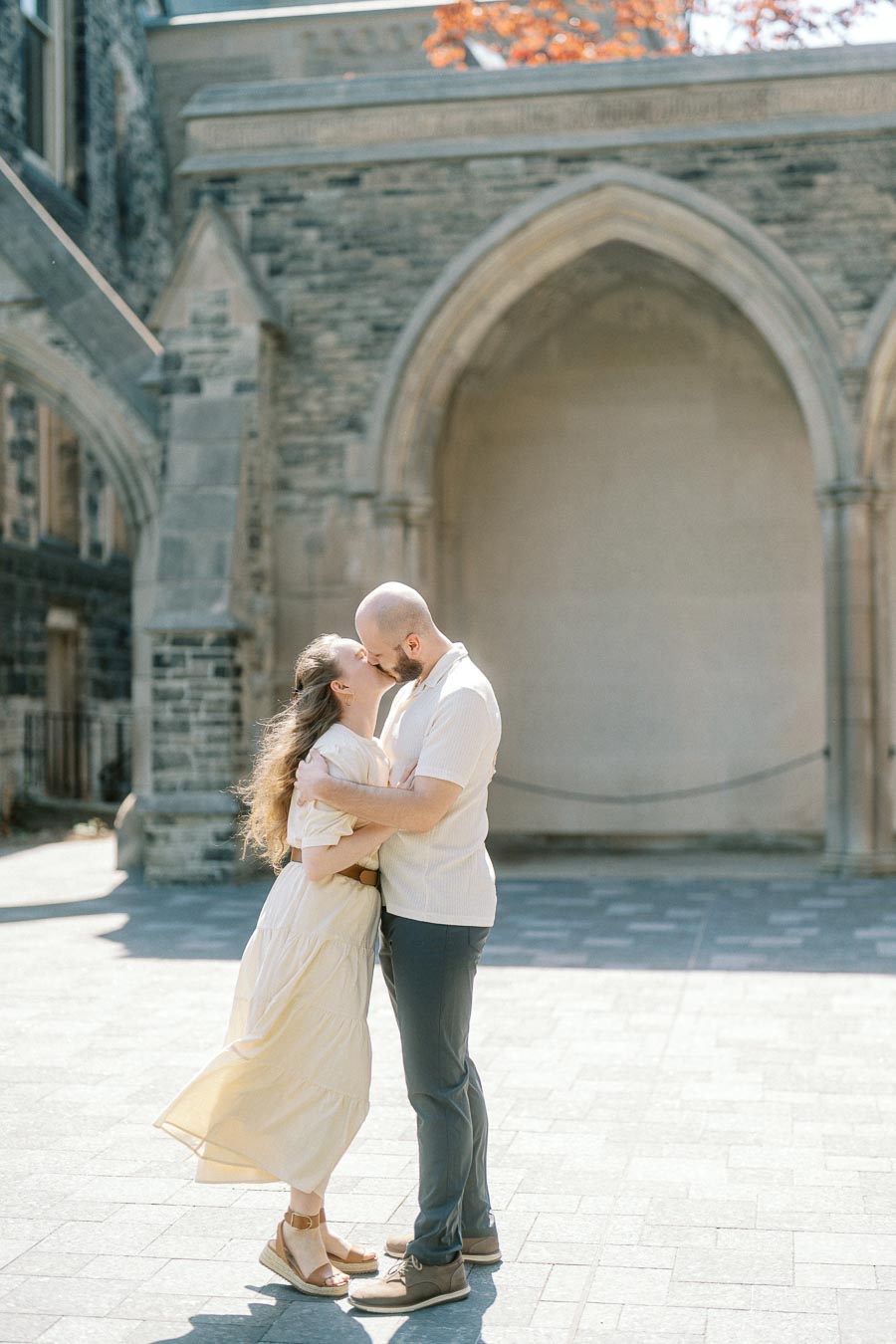 Engaged couple shares a kiss in bright sunlight under a historic stone archway at the University of Toronto campus in spring.