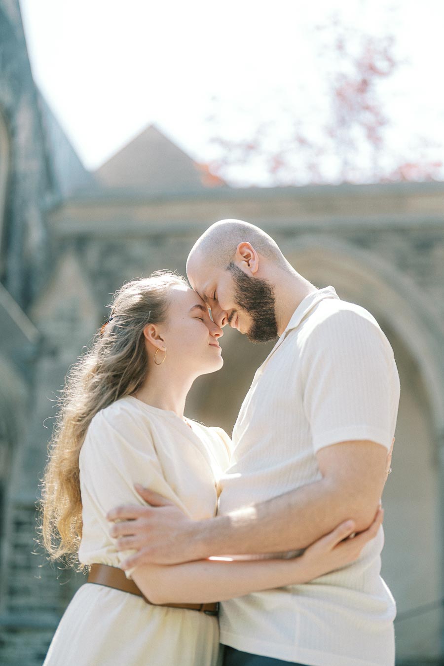 Couple shares a gentle forehead touch in soft sunlight, surrounded by historic stone arches and spring foliage at University of Toronto.
