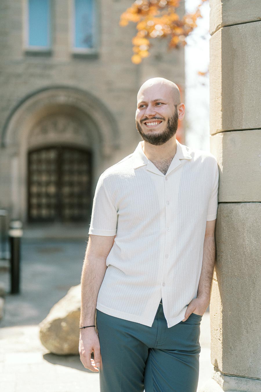 Hayden smiles warmly while leaning on a stone column with historic university architecture and spring foliage in the background.