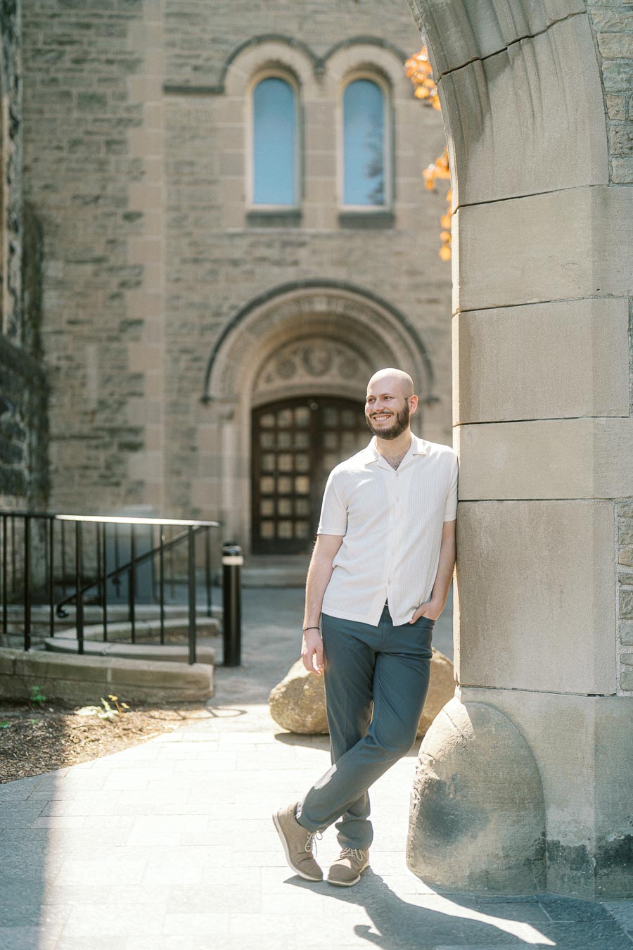 Hayden leans casually against a stone archway at the University of Toronto, smiling in soft spring sunlight with historic architecture behind.