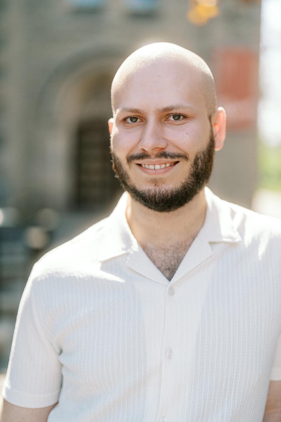 A man in a light shirt smiles warmly in front of a sunlit stone archway at the University of Toronto campus.