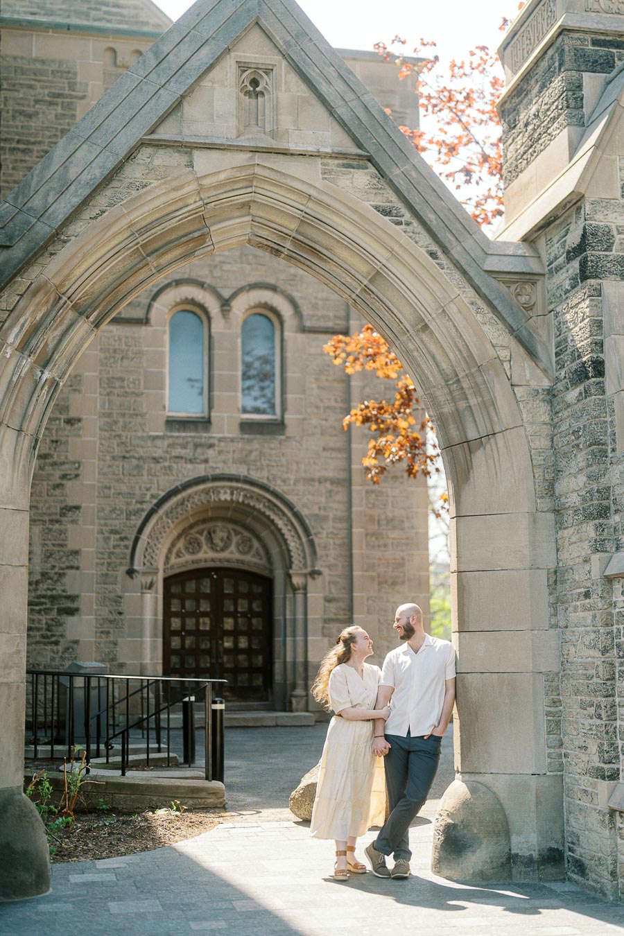 Couple smiles and holds hands under a sunlit stone archway at the University of Toronto, surrounded by historic architecture and spring foliage.