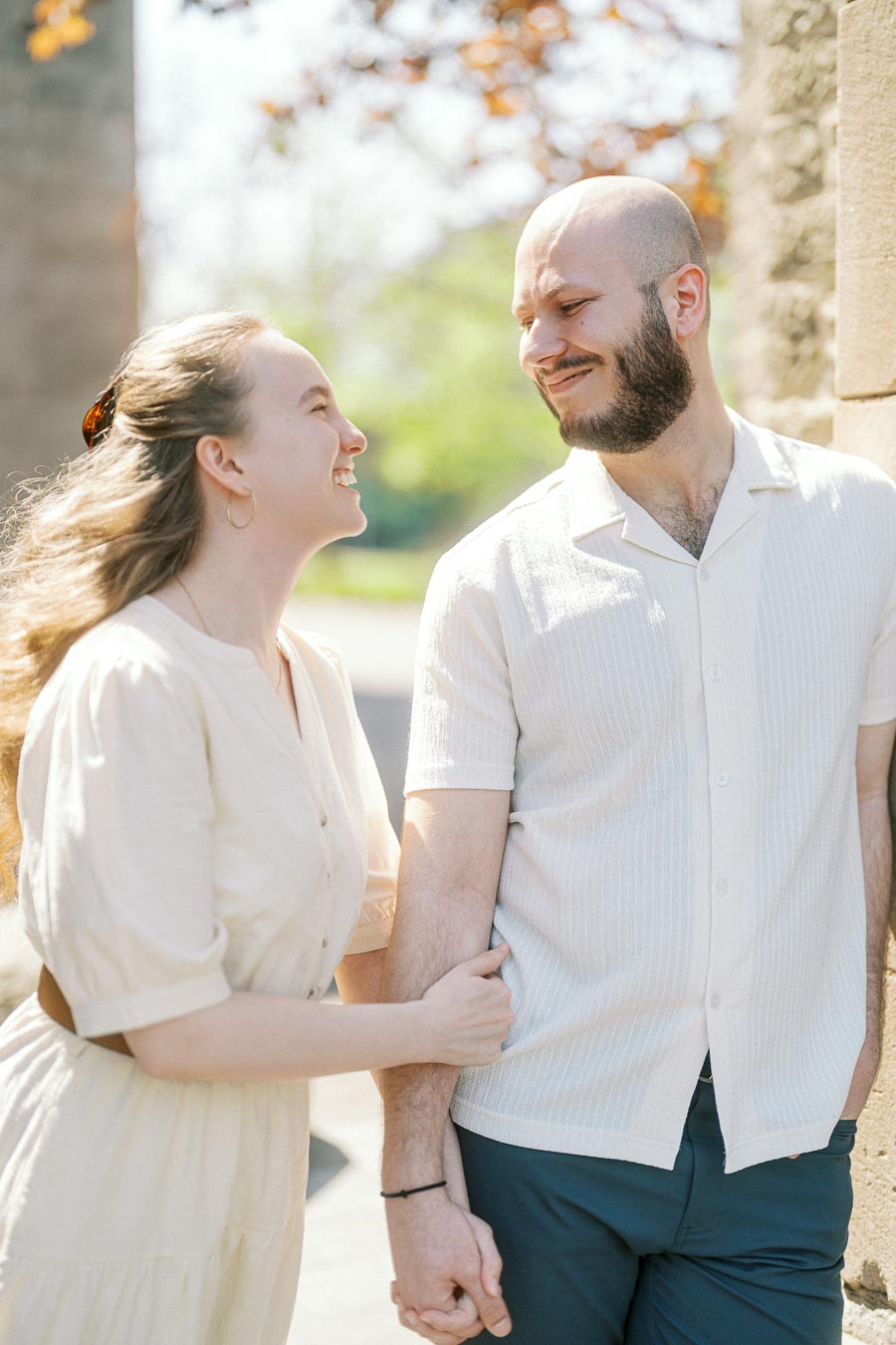 Couple smiling at each other while holding hands in natural light beside a historic stone wall at University of Toronto in spring.