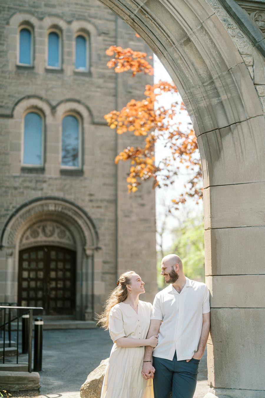 A couple stands hand in hand under a stone arch at the University of Toronto, smiling at each other with sunlight and blooming trees in the background.