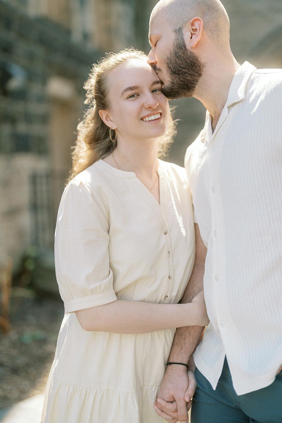 Smiling woman in a cream dress holds hands with her partner as he kisses her forehead, standing in soft spring sunlight at University of Toronto.